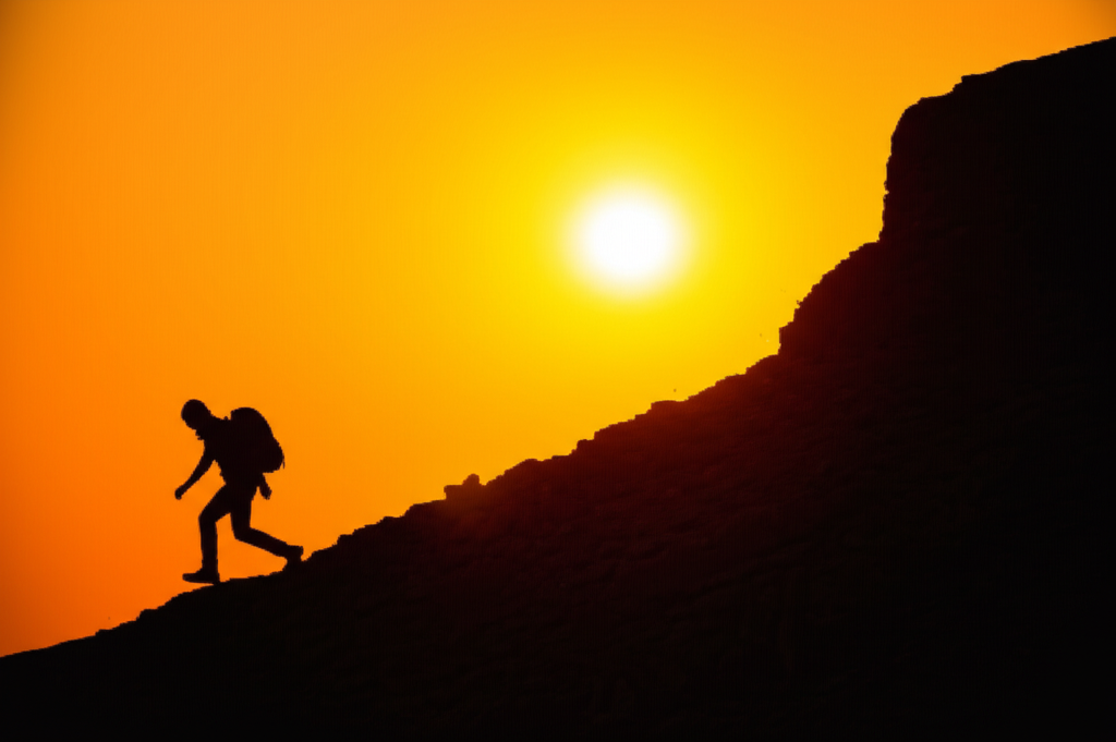 A person silhouetted against a bright, setting sun, climbing a steep, rocky mountain path.